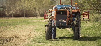 Zdjęcie przedstawia rolnika stojącego przy swym traktorze. Obok niego pole, na nim owoce pracy wzrastają, wokół porastających scenę drzew.