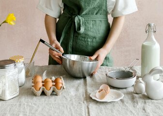 Obraz przedstawia ręce wyrabiającej ciasto osoby. Na pierwszym planie znajduje się stół zastawiony składnikami.