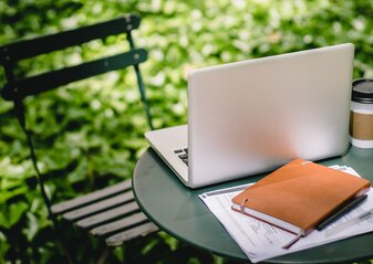 Zdjęcie przedstawia laptopa i notatnik na stole w otoczeniu zieleni ogrodu.