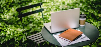 Zdjęcie przedstawia laptopa i notatnik na stole w otoczeniu zieleni ogrodu.