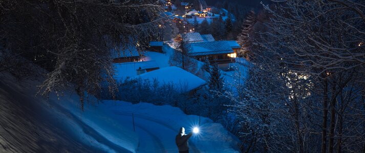 Zdjęcie przedstawia krajobraz górski osnuty mrokiem. Na pierwszym planie jest osoba z latarką, oświetlająca sobie drogę w dół, prowadzącą do niżej położonej, oświetlonej wsi. 