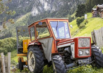 Na zdjęciu znajduje się traktor stojący przed gospodarstwem, zaraz przy górskiej drodze.