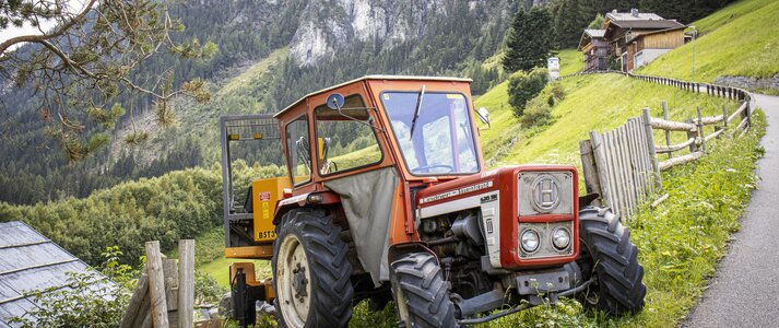Na zdjęciu znajduje się traktor stojący przed gospodarstwem, zaraz przy górskiej drodze.