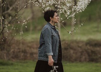 Zdjęcie przedstawia kobietę stojącą pod drzewem, trzymającą gałąź kwitnącą.