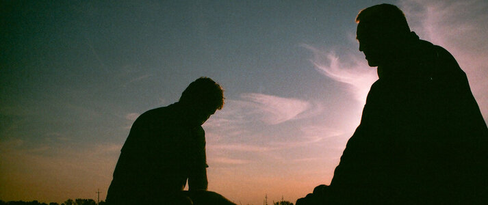 Zdjęcie przedstawia sylwetki dwóch pochylonych mężczyzn na polu, po pracy, o zachodzi słońca.
