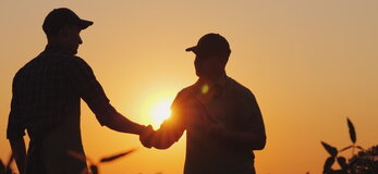 Zdjęcie przedstawia dwóch rolników podających sobie rękę na polu, o zachodzie słońca.