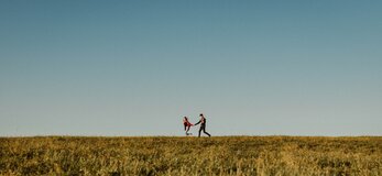 Zdjęcie przedstawia dwójkę osób biegnących w lewą stronę, trzymających się za ręce, znajdujących się na linii horyzontu.