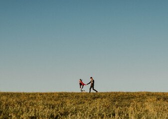Zdjęcie przedstawia dwójkę osób biegnących w lewą stronę, trzymających się za ręce, znajdujących się na linii horyzontu.