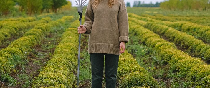 Zdjęcie przedstawia sylwetkę kobiety stojącej na środku pola, trzymającej grabie.