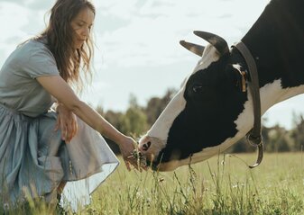 Obraz przedstawia dziewczynę na łące, karmiącą z ręki krowę.