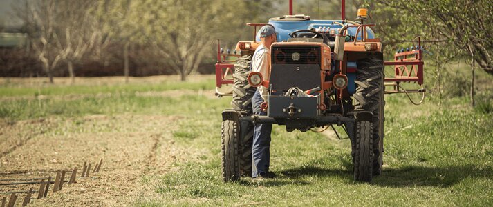 Zdjęcie przedstawia rolnika stojącego przy swym traktorze. Obok niego pole, na nim owoce pracy wzrastają, wokół porastających scenę drzew.