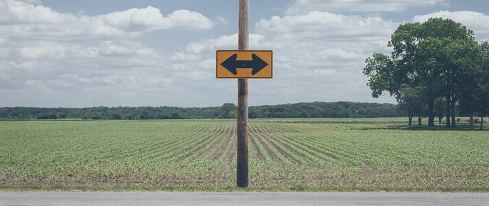 Zdjecie przedstawia znak ze strzalkami w dwoch przeciwnych kierunkach, znajdujący się na pustym polu na wsi.